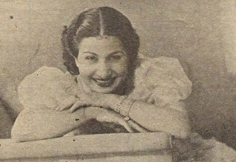 Sepia magazine photograph of a smiling Umm Kulthum leaning on a railing in a puff-sleeved dress, resting her folded arms.