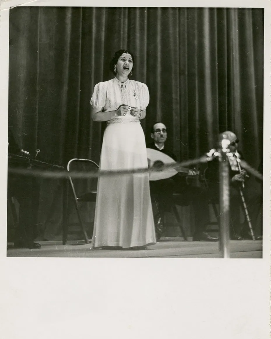 Black-and-white stage photograph of Umm Kulthum singing in a long white gown before a draped curtain, with oud and violin players seated behind her.