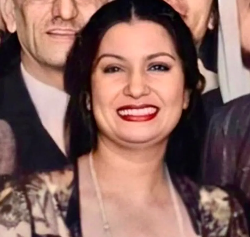 Colorized close-up of a smiling Umm Kulthum with dark hair and red lipstick amid a crowd of men in suits at an event.
