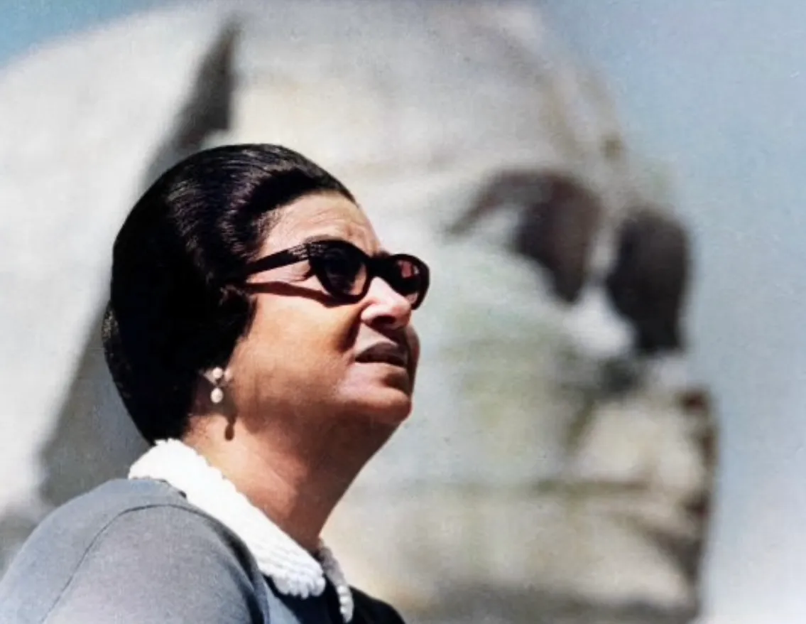 Color profile photograph of Umm Kulthum wearing dark sunglasses, pearl earrings and a grey collared dress, gazing upward outdoors.