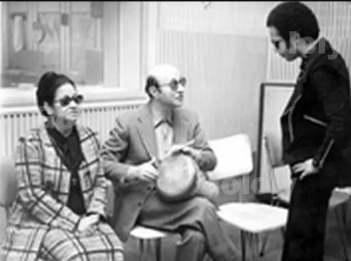 Black-and-white backstage photograph of Umm Kulthum in sunglasses seated beside composer Mohammed Abdel Wahab holding a tambourine, with a young man standing.