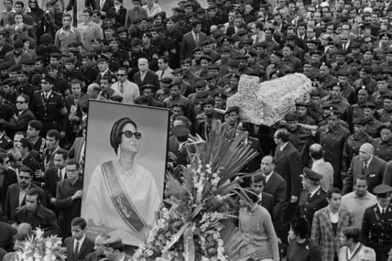 Black-and-white photograph of a vast crowd carrying Umm Kulthum's flower-draped coffin and a large framed portrait of her through the street.