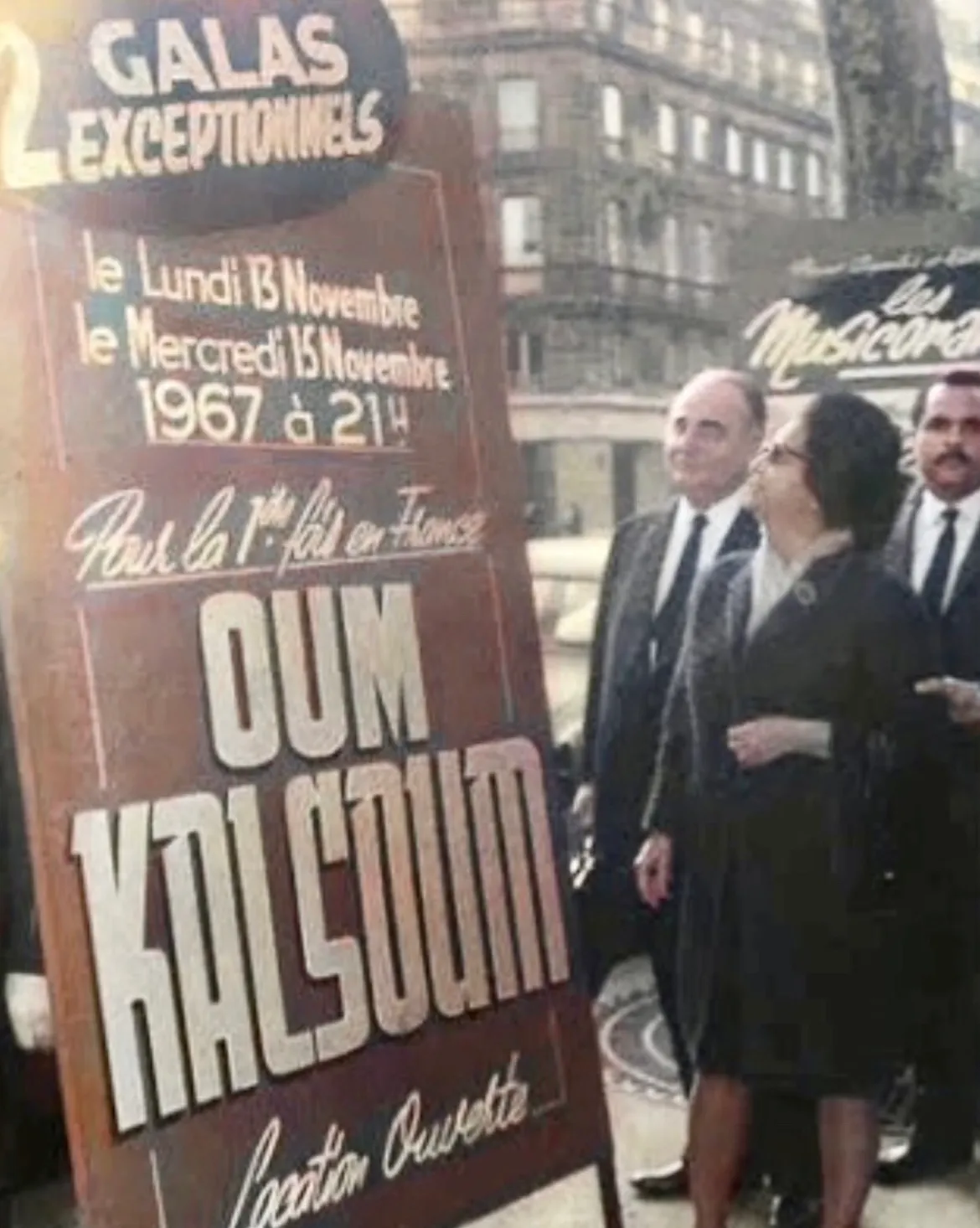 Umm Kulthum in a dark coat and sunglasses stands beside men in suits before a Paris billboard advertising her 1967 Olympia galas.