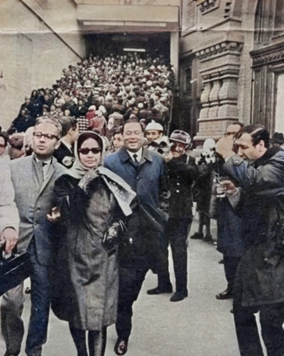 Colorized photograph of Umm Kulthum in a headscarf, sunglasses and fur-trimmed coat walking through a crowd with companions and photographers.