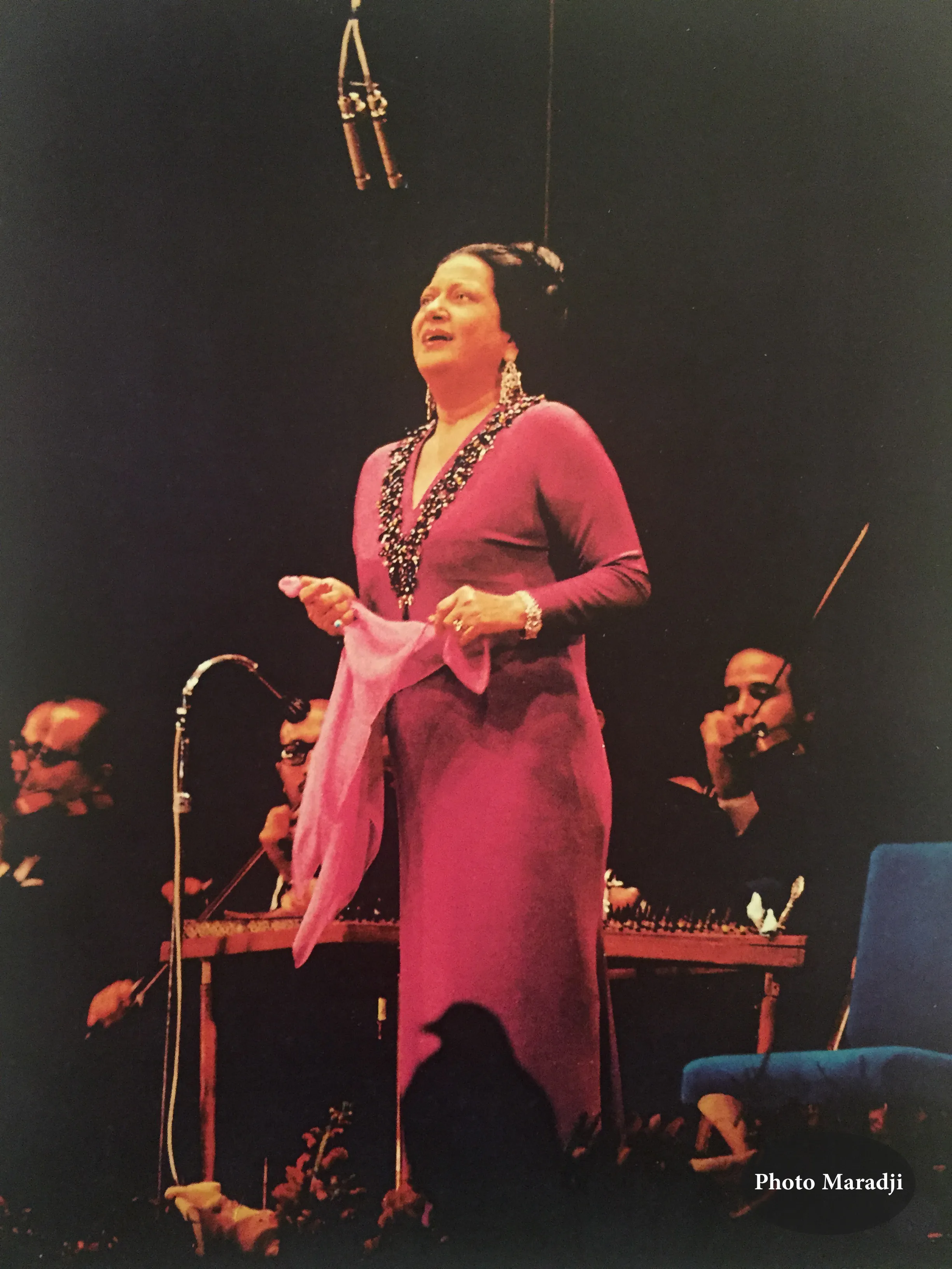 Color concert photograph of Umm Kulthum in a magenta gown holding a scarf and singing on a darkened stage, backed by her orchestra.