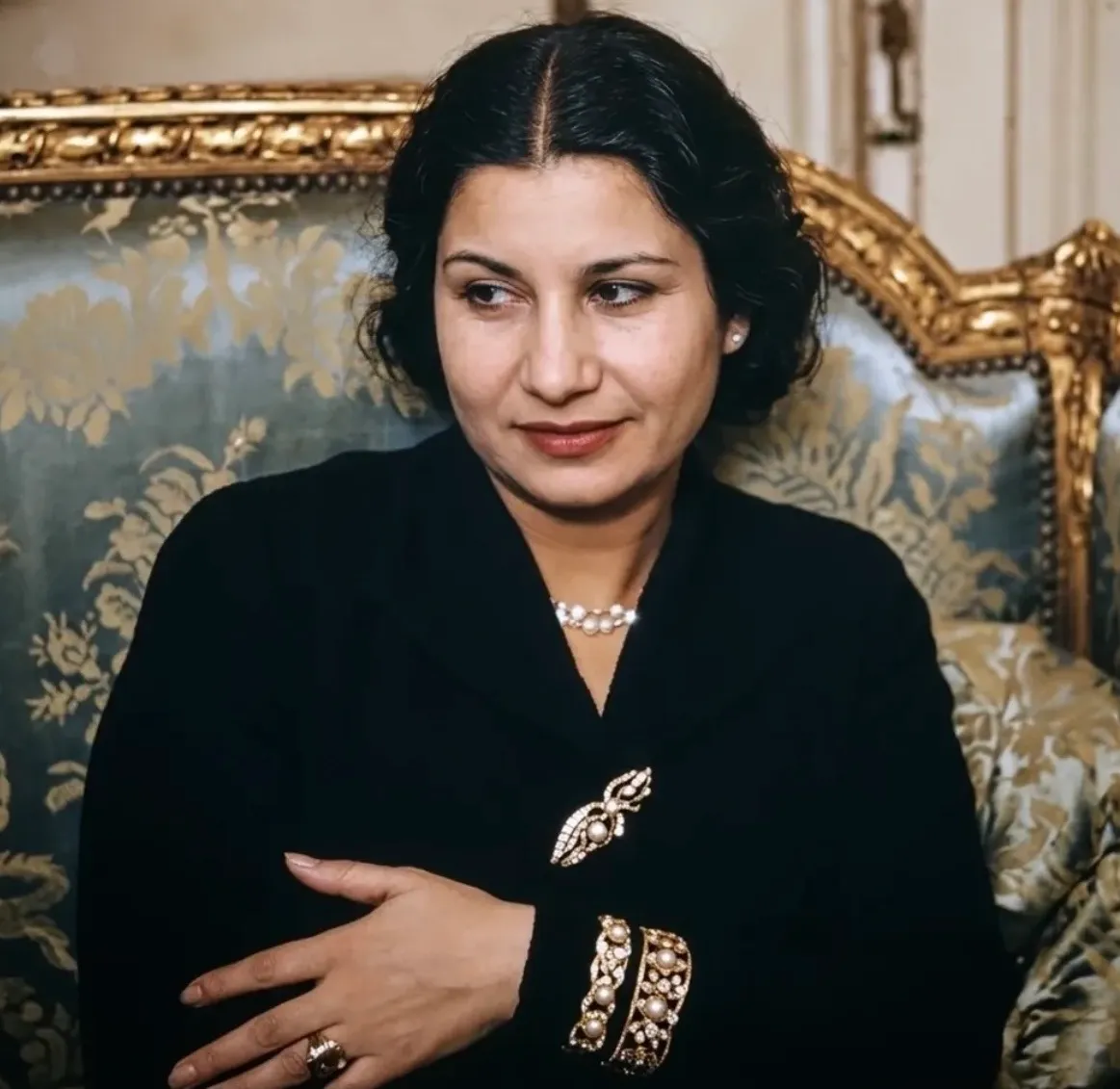 Colorized portrait of Umm Kulthum in a black dress with a pearl necklace and jeweled brooch, seated on a gilded brocade sofa looking aside.