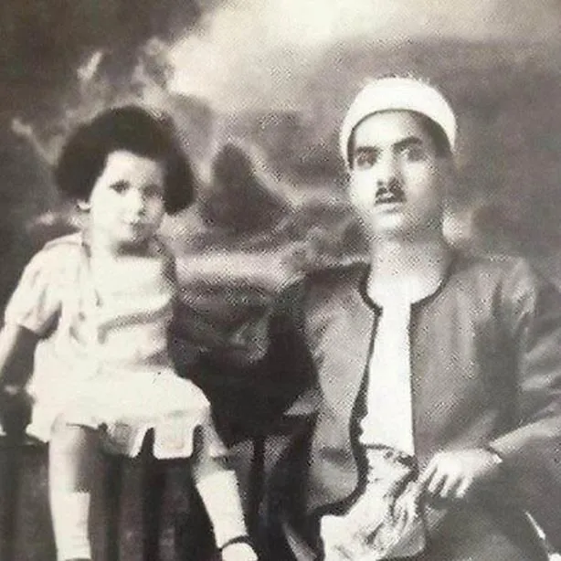 Aged studio photograph of Umm Kulthum as a small child seated beside her father, a mustached man in a turban and traditional robe.